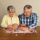 Two older adults engaged in assembling the Springtime Goldfinches jigsaw puzzle from Cobble Hill. The colorful puzzle features vibrant flowers and goldfinches, with pieces scattered around them on a wooden table.