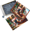 A Cobble Hill jigsaw puzzle titled 'Chez Michelle'. The box is open, revealing numerous gray puzzle pieces, with one piece displayed outside the box. The puzzle image shows a charming café scene with colorful chairs, tables, and vibrant flowers, set on a cobblestone street.