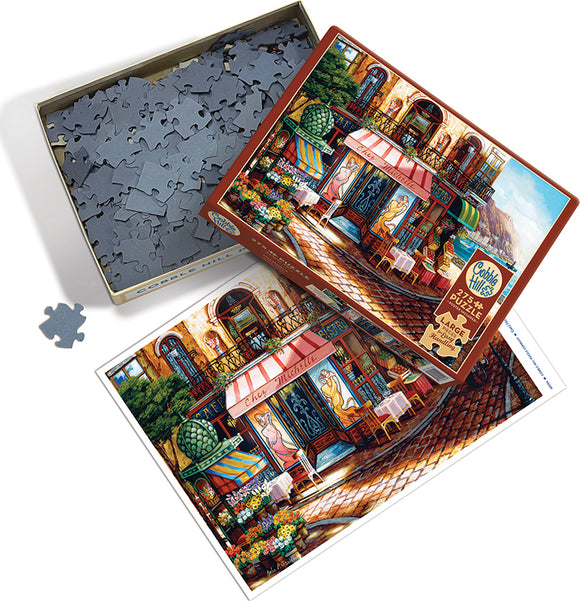 A Cobble Hill jigsaw puzzle titled 'Chez Michelle'. The box is open, revealing numerous gray puzzle pieces, with one piece displayed outside the box. The puzzle image shows a charming café scene with colorful chairs, tables, and vibrant flowers, set on a cobblestone street.