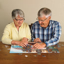 An elderly couple engaged in assembling the Chez Michelle jigsaw puzzle made by Cobble Hill. They are seated at a wooden table with scattered puzzle pieces around them, smiling and concentrating on their task.
