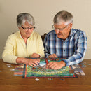 A senior couple enjoys assembling the Tree of Life Stained Glass jigsaw puzzle made by Cobble Hill on a wooden table. The puzzle features a vibrant stained glass design depicting a tree surrounded by lush greenery, with several pieces scattered around them. The man, wearing a plaid shirt, and the woman, in a yellow sweater, are focused and smiling as they connect puzzle pieces together.