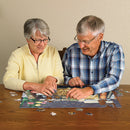 An elderly couple assembling a jigsaw puzzle together on a wooden table. The woman wears glasses and a light yellow sweater, while the man wears a plaid shirt. Puzzle pieces are scattered around them.