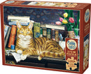 Cobble Hill 275-piece jigsaw puzzle featuring a relaxed orange tabby cat lounging on a desk surrounded by books, flowers, and a decorative background. The design showcases a cozy reading nook with various titles visible on the bookshelf.