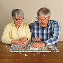 An elderly couple enjoying a jigsaw puzzle together at a wooden table. The puzzle features a colorful design with flowers and cacti. They are focused on fitting pieces together, surrounded by additional puzzle pieces. The scene conveys a sense of joy and companionship.