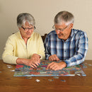 An elderly couple working on the Cobble Hill 'Trolley Station' jigsaw puzzle together at a wooden table. The puzzle pieces are colorful, depicting a charming street scene with trees and buildings. The couple shows joy and concentration as they piece together the puzzle, surrounded by scattered pieces.