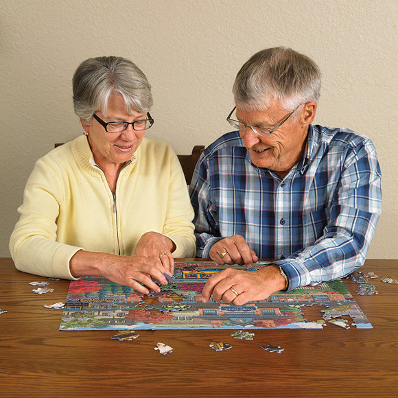 An elderly couple working on the Cobble Hill 'Trolley Station' jigsaw puzzle together at a wooden table. The puzzle pieces are colorful, depicting a charming street scene with trees and buildings. The couple shows joy and concentration as they piece together the puzzle, surrounded by scattered pieces.