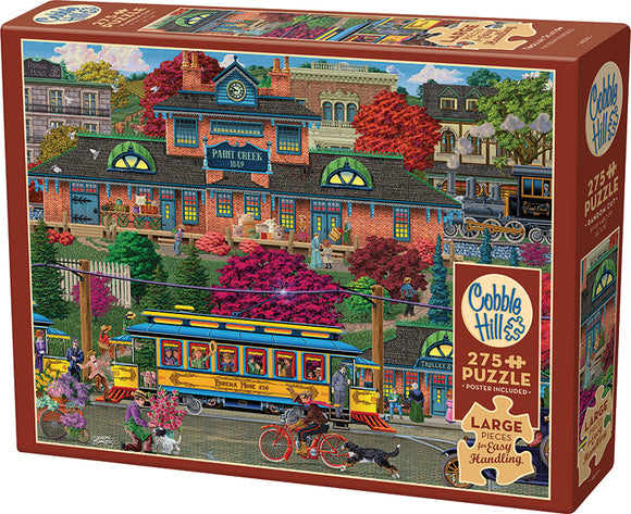 Cobble Hill Trolley Station jigsaw puzzle featuring a vibrant, detailed illustration of a historic trolley station scene with people interacting, colorful trees, flowers, and vintage vehicles. The box includes a 275-piece puzzle and a poster for reference.