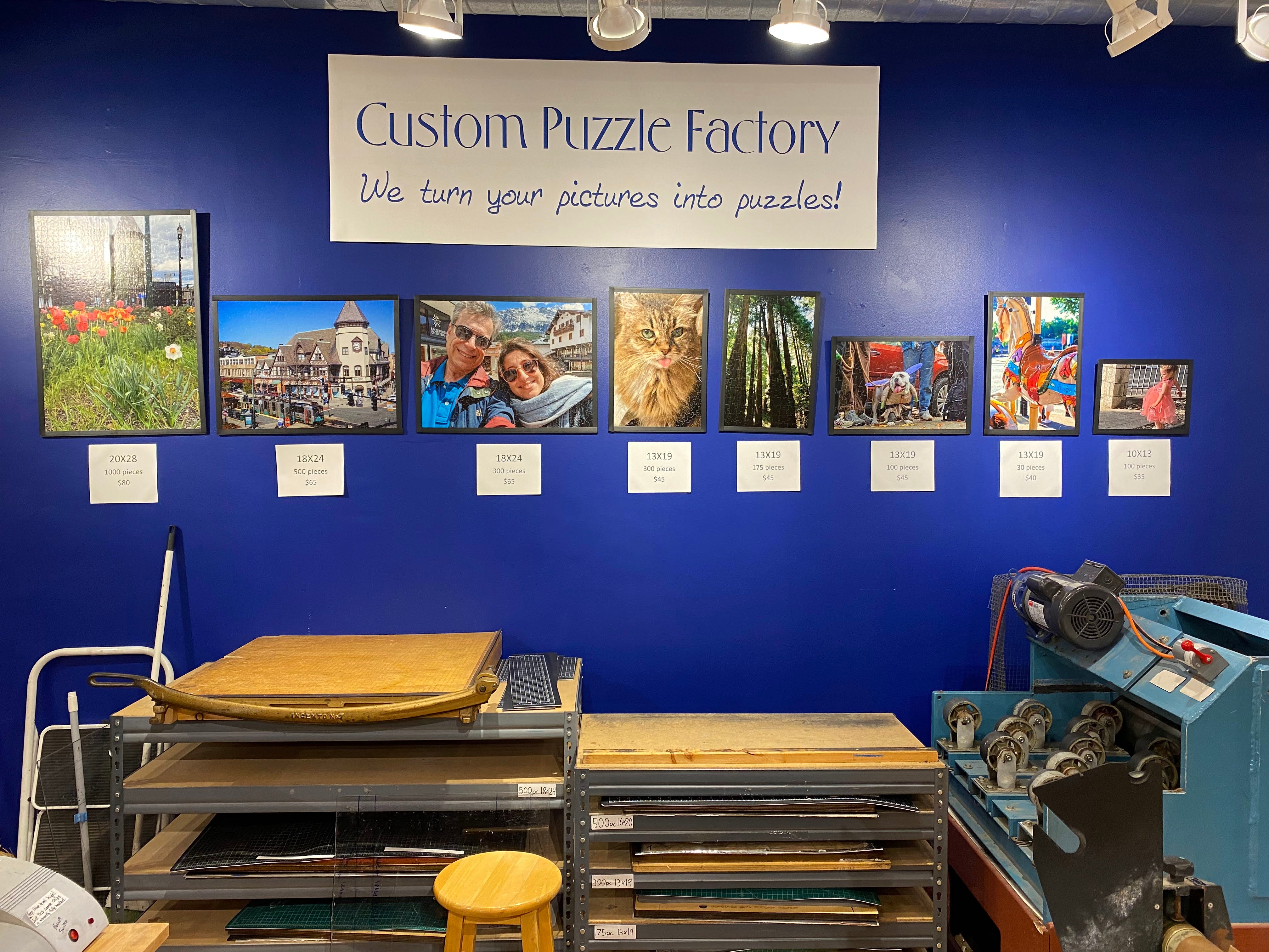 Custom Jigsaw puzzle display at Eureka Puzzles, showcasing various completed puzzles with images of flowers, landscapes, pets, and people. The wall features a sign that reads 'Custom Puzzle Factory' and descriptions of puzzle sizes and prices beneath each image.