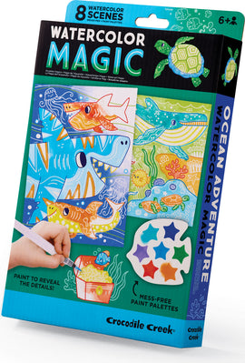 The Watercolor Magic Ocean Adventure kit by Crocodile Creek features eight watercolor scenes, each illustrating vibrant ocean life with images of sharks, turtles, and colorful fish. The kit includes a brush pen and mess-free paint palettes, designed for ages 6 and up. A hand holds the brush pen over a scene, demonstrating the painting process to reveal hidden details.