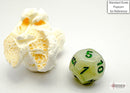 A close-up of a polyhedral die made of green and dark green marbled material, showing the numbers 1 to 10. Next to the die is a small piece of popcorn for scale reference, highlighting the size of the die. The background is a neutral tone, emphasizing the vivid colors of the die.