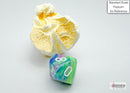 Festive mini poly waterlily die by Chessex, featuring a vibrant blend of green, blue, and purple colors with white numbers. The die is shown alongside a piece of popcorn for scale.