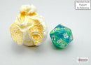 A close-up image of a Festive mini poly waterlily die beside a piece of popcorn for scale. The die features vibrant blue and green swirls with white numbers, showcasing its unique design and craftsmanship by Chessex. It is perfect for gaming enthusiasts looking to add a colorful touch to their collection.