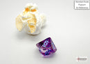 A close-up image of the Nebula mini poly nocturn blue game dice by Chessex, featuring a striking purple and blue design with white numbers. A piece of popcorn is included for size reference.