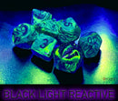 A set of six festive polyhedral dice in shades of green and silver, featuring numbers highlighted in a contrasting color. The dice are displayed under black light, enhancing their vibrant colors and creating a luminous effect, perfect for tabletop games.