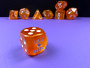 A vibrant Borealis Blood Orange and White Luminary 7 Piece Dice Set by Chessex. The set features a variety of polyhedral dice with a glossy orange finish and white numbers. The largest die displays 'LAB DICE' engraved, standing out against a purple surface, with additional dice featuring numbers arranged artistically in the background.