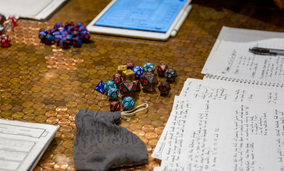 A tabletop scene featuring a variety of colorful polyhedral dice scattered among notes and a tablet. The tabletop surface is made of shiny copper pennies, creating a warm background. The dice colors include blue, red, yellow, and brown, which are often used in tabletop role-playing games. There's also a small gray pouch and several sheets of handwritten notes, detailing game mechanics or character information for an engaging RPG experience.