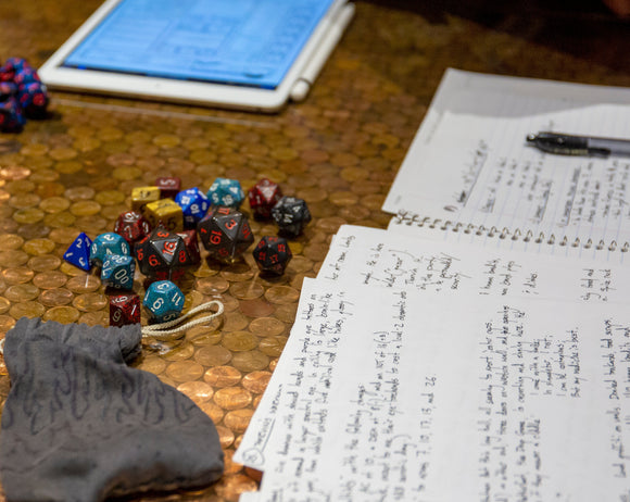A tabletop setup for a Dungeons & Dragons game featuring a variety of colorful polyhedral dice scattered over a copper penny surface. Nearby, there are notebooks filled with handwritten notes and an electronic tablet displaying a game sheet, adding to the immersive role-playing atmosphere.