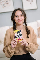 A young girl with braided hair is smiling while holding the card game 'Taco Cat Goat Cheese Pizza' by Dolphin Hat Games. She is wearing a cozy brown sweater and sitting in a softly lit room, with decorative art in the background.
