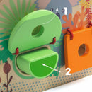 An educational toy featuring colorful wooden shapes designed for children. The image shows a green half-moon shape being positioned into a matching slot on a vibrant puzzle board, complete with playful illustrations of flora and fauna in the background.