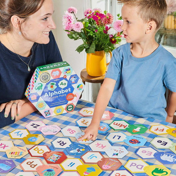 A mother and son engage with the Eeboo Alphabet Puzzle Pairs on a checkered tablecloth. The colorful puzzle pieces feature letters and playful illustrations, while the packaging showcases vibrant designs. A yellow vase filled with flowers adds charm to the scene.