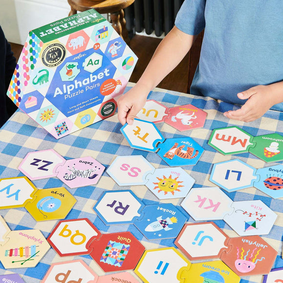 A young child plays with the Eeboo Alphabet Puzzle Pairs, which features colorful hexagonal pieces showcasing various letters and associated illustrations. The puzzle pieces include images of animals and objects, designed to engage children while they learn the alphabet. The packaging is bright and eye-catching, emphasizing educational fun and creativity.