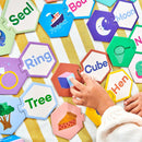 A hand is reaching for a colorful hexagon puzzle piece featuring the word 'Cube.' The puzzle includes various pieces with illustrations and words, such as 'Seal,' 'Tree,' and 'Hen.' The pieces are arranged on a striped background, showcasing bright visuals and engaging design, ideal for learning and play.