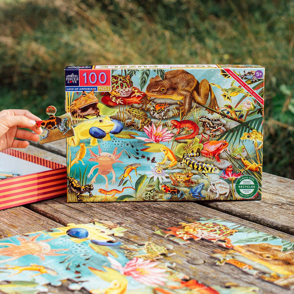 The Love of Amphibians 100-piece jigsaw puzzle by Eeboo showcases vibrant illustrations of various amphibian species, set against a lush, aquatic backdrop. A hand holds a puzzle piece, and several completed pieces are scattered across a rustic wooden table, emphasizing the engaging and educational experience of piecing together this colorful scene.