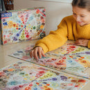 A joyful child wearing a fluffy yellow jacket is putting together a colorful 100-piece jigsaw puzzle featuring various crystals and gems. The puzzle box, displayed nearby, shows intricate illustrations of the gems, including labels for each type. The table is covered with pieces of the puzzle, showcasing vibrant colors and unique shapes, perfect for engaging young minds in creative play.