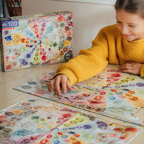 A joyful child wearing a fluffy yellow jacket is putting together a colorful 100-piece jigsaw puzzle featuring various crystals and gems. The puzzle box, displayed nearby, shows intricate illustrations of the gems, including labels for each type. The table is covered with pieces of the puzzle, showcasing vibrant colors and unique shapes, perfect for engaging young minds in creative play.