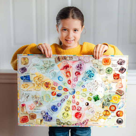 A child holds a completed 100-piece jigsaw puzzle titled 'Love of Crystal & Gems' by Eeboo. The puzzle features a vibrant display of various crystals and gems, each labeled with their names, arranged in a colorful circular pattern.