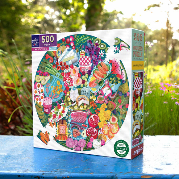 Eeboo Charcuterie 500-piece round jigsaw puzzle displayed outdoors. The colorful box features a vibrant assortment of charcuterie items, including fruits, meats, and cheese, set against a natural backdrop. This beautifully illustrated puzzle invites creativity and is made from recycled materials.