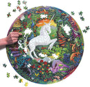 A vibrant round jigsaw puzzle titled 'Unicorn Garden' by Eeboo, featuring a white unicorn surrounded by colorful flora and fauna. A hand is shown placing a piece in the puzzle, with several pieces scattered nearby.