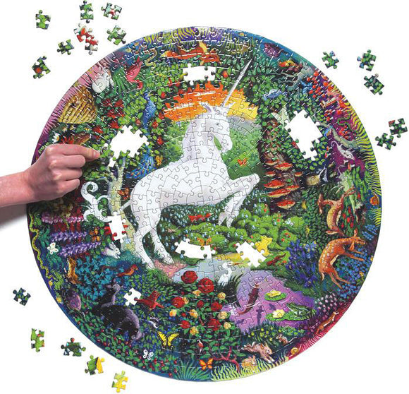A vibrant round jigsaw puzzle titled 'Unicorn Garden' by Eeboo, featuring a white unicorn surrounded by colorful flora and fauna. A hand is shown placing a piece in the puzzle, with several pieces scattered nearby.
