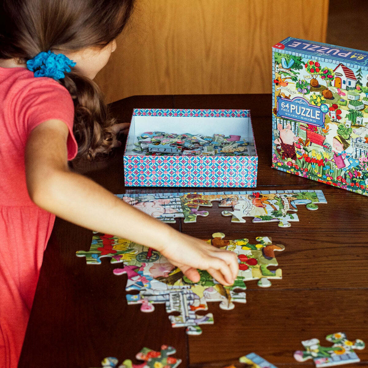 A child working on the Eeboo 'Growing a Garden' jigsaw puzzle on a wooden table. The colorful puzzle pieces are scattered around, and a decorative box containing more pieces is open beside them. The puzzle features vibrant illustrations of a garden scene, making it engaging for kids.