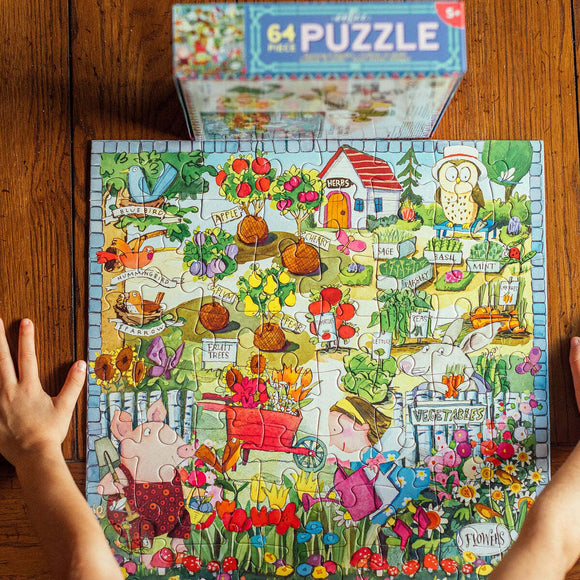 A colorful 64-piece jigsaw puzzle titled 'Growing a Garden' by Eeboo. The puzzle depicts a vibrant garden scene including various animals and plant labels, with a child watering flowers and a pig in a wheelbarrow. The puzzle pieces are partially assembled on a wooden table, with a box of the puzzle visible in the background.