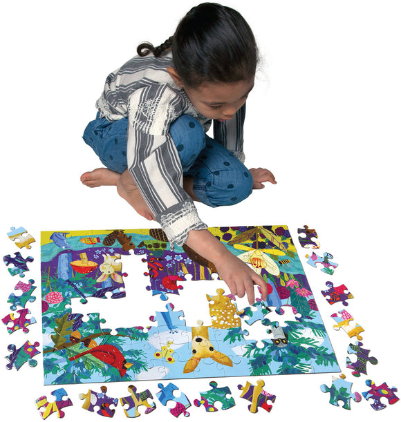 A young girl with braided hair is focused on assembling the 'Life on Earth' 100 piece jigsaw puzzle. She is sitting on the floor and has a variety of colorful puzzle pieces spread out around her, working to complete a vibrant image of nature featuring various animals and plants.