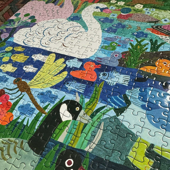 A colorful jigsaw puzzle featuring a vibrant scene of various ducks, swans, and fish swimming in a pond. The illustration includes a white swan, orange ducklings, and blue and yellow fish among lush greens and aquatic plants, showcasing a playful and engaging design suitable for all ages.