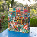 The English Green Market 1000-piece jigsaw puzzle by Eeboo features a vibrant illustration of a bustling market scene, showcasing colorful stalls filled with flowers, fruits, and baked goods. The box art includes quaint shops with awnings, cheerful vendors, and lively characters, all set in a picturesque outdoor setting. The product emphasizes eco-friendly materials with a 'recycled' badge.