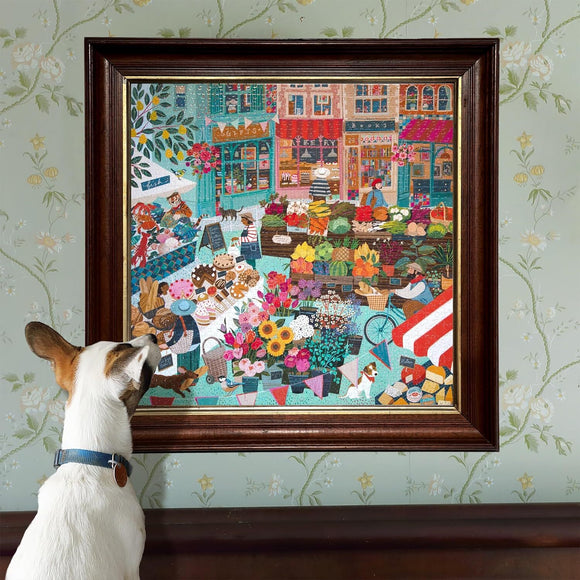 A framed jigsaw puzzle depicting a vibrant English market scene filled with colorful flowers, fruits, and baked goods. A small dog curiously gazes at the artwork, enhancing the cozy atmosphere of a home decorated with floral wallpaper.