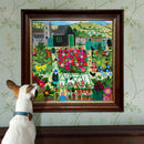 A playful dog looks at a framed jigsaw puzzle featuring a vibrant garden scene filled with colorful flowers, plants, greenhouses, and a quaint house, set against a backdrop of rolling hills.