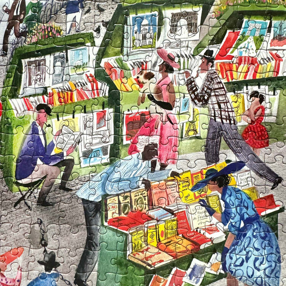 A vibrant jigsaw puzzle depicting a Parisian bookseller scene. The image features people browsing colorful book stalls along a street in Paris, showcasing lively details like books of various sizes and characters immersed in reading and walking. The artwork highlights the cultural charm of Paris with its iconic ambiance.