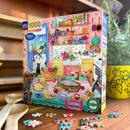 Eeboo's Pink Kitchen 1000-piece jigsaw puzzle featuring a vibrant and colorful kitchen scene. The box showcases a beautifully illustrated kitchen filled with a variety of items, including fruits, vegetables, kitchenware, and playful pets. The setting includes pink walls, a charming yellow sink, and a view of a scenic outdoor patio, creating a lively and warm atmosphere perfect for puzzle enthusiasts.