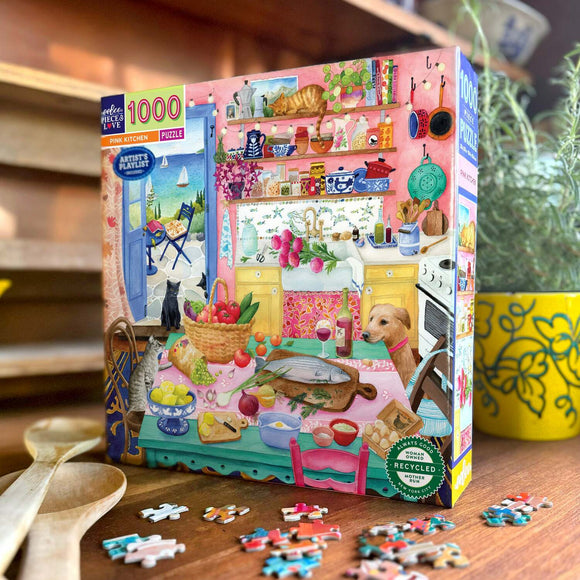 Eeboo's Pink Kitchen 1000-piece jigsaw puzzle featuring a vibrant and colorful kitchen scene. The box showcases a beautifully illustrated kitchen filled with a variety of items, including fruits, vegetables, kitchenware, and playful pets. The setting includes pink walls, a charming yellow sink, and a view of a scenic outdoor patio, creating a lively and warm atmosphere perfect for puzzle enthusiasts.