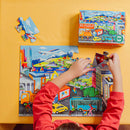 A child with a red shirt is assembling a 20-piece jigsaw puzzle featuring various vehicles including cars, boats, and planes. The colorful puzzle depicts a vibrant city scene. The puzzle box can be seen in the background, showcasing the Eeboo brand and its eco-friendly material.