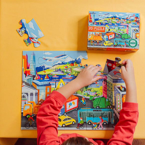 A child with a red shirt is assembling a 20-piece jigsaw puzzle featuring various vehicles including cars, boats, and planes. The colorful puzzle depicts a vibrant city scene. The puzzle box can be seen in the background, showcasing the Eeboo brand and its eco-friendly material.