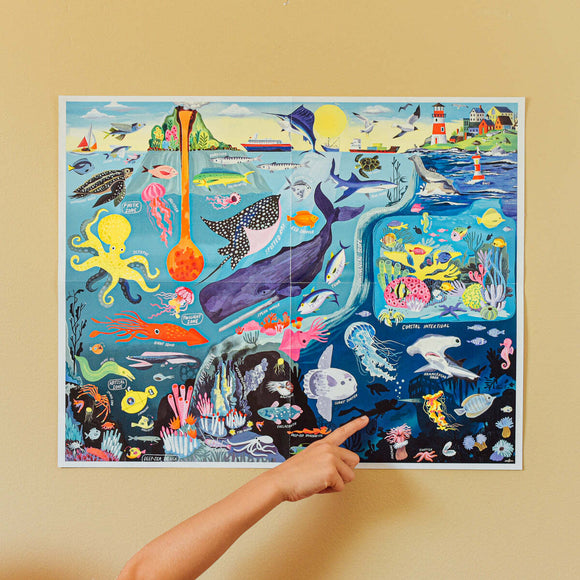 A vibrant 48-piece jigsaw puzzle titled 'Within the Sea' by Eeboo. The puzzle features an illustrated underwater scene filled with colorful sea creatures, coral reefs, and other marine life. A hand is pointing at a specific area of the puzzle, showcasing the intricate details and educational aspects of the design.
