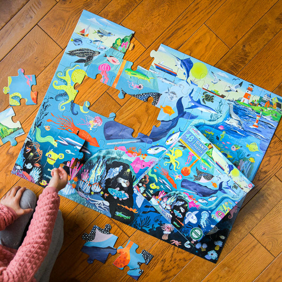 A vibrant 48-piece jigsaw puzzle titled 'Within the Sea' by Eeboo, featuring a colorful underwater scene filled with sea creatures and plants. A child's hand is seen fitting a piece into the puzzle on a wooden floor.