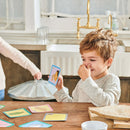 A cheerful young boy with light brown hair sits at a wooden table, holding a card from the Scavenger Hunt Game by Eeboo. He has a playful expression, pinching his nose as he looks at the card. Colorful game cards are scattered on the table, creating an engaging indoor game atmosphere.
