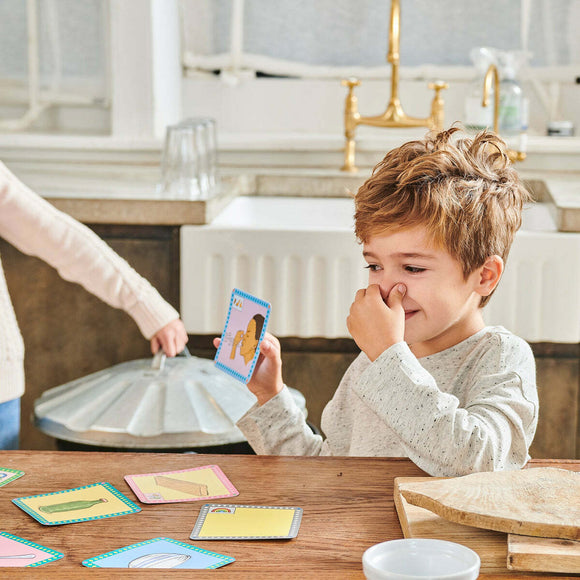 A cheerful young boy with light brown hair sits at a wooden table, holding a card from the Scavenger Hunt Game by Eeboo. He has a playful expression, pinching his nose as he looks at the card. Colorful game cards are scattered on the table, creating an engaging indoor game atmosphere.