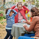 Two children enjoying a scavenger hunt game outdoors, excitedly reacting while a woman holds up a card at a table covered with a checkered cloth, surrounded by greenery.